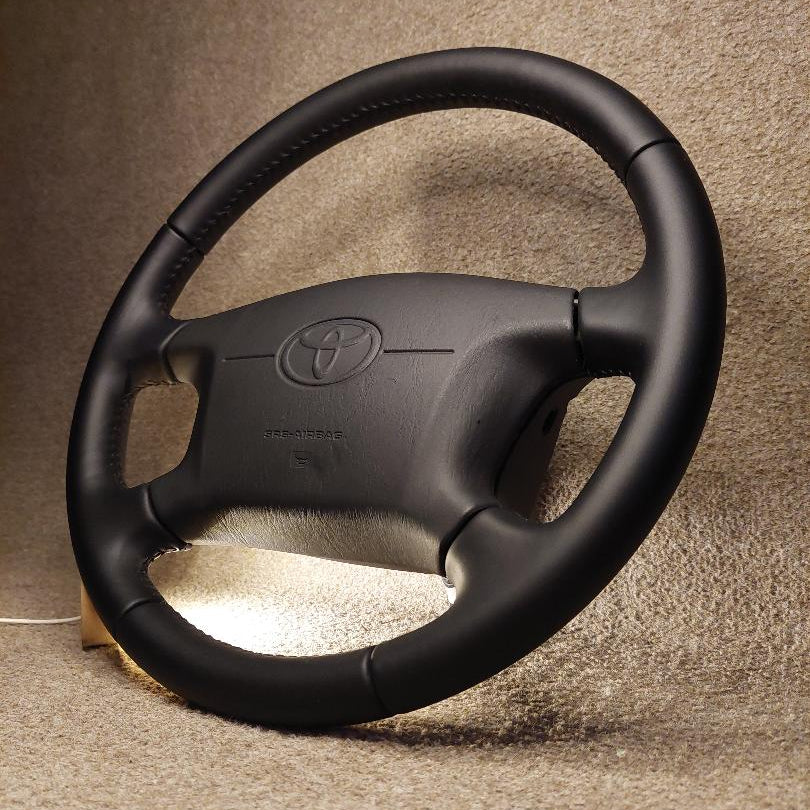 A black leather-wrapped Toyota steering wheel shown from a front-facing angle, resting on a textured beige carpeted floor.