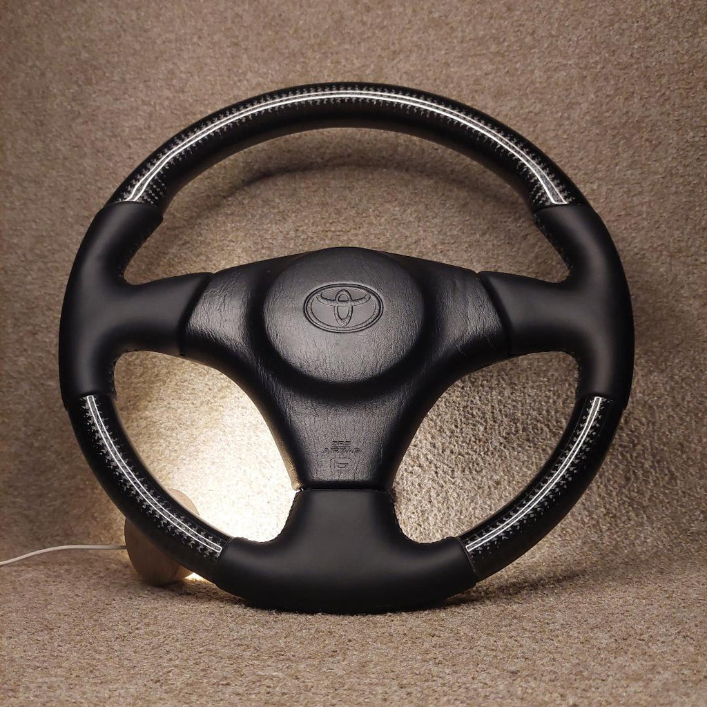 A black leather steering wheel cover with white contrast stitching and a central Toyota emblem, shown on a steering wheel against a textured beige background.