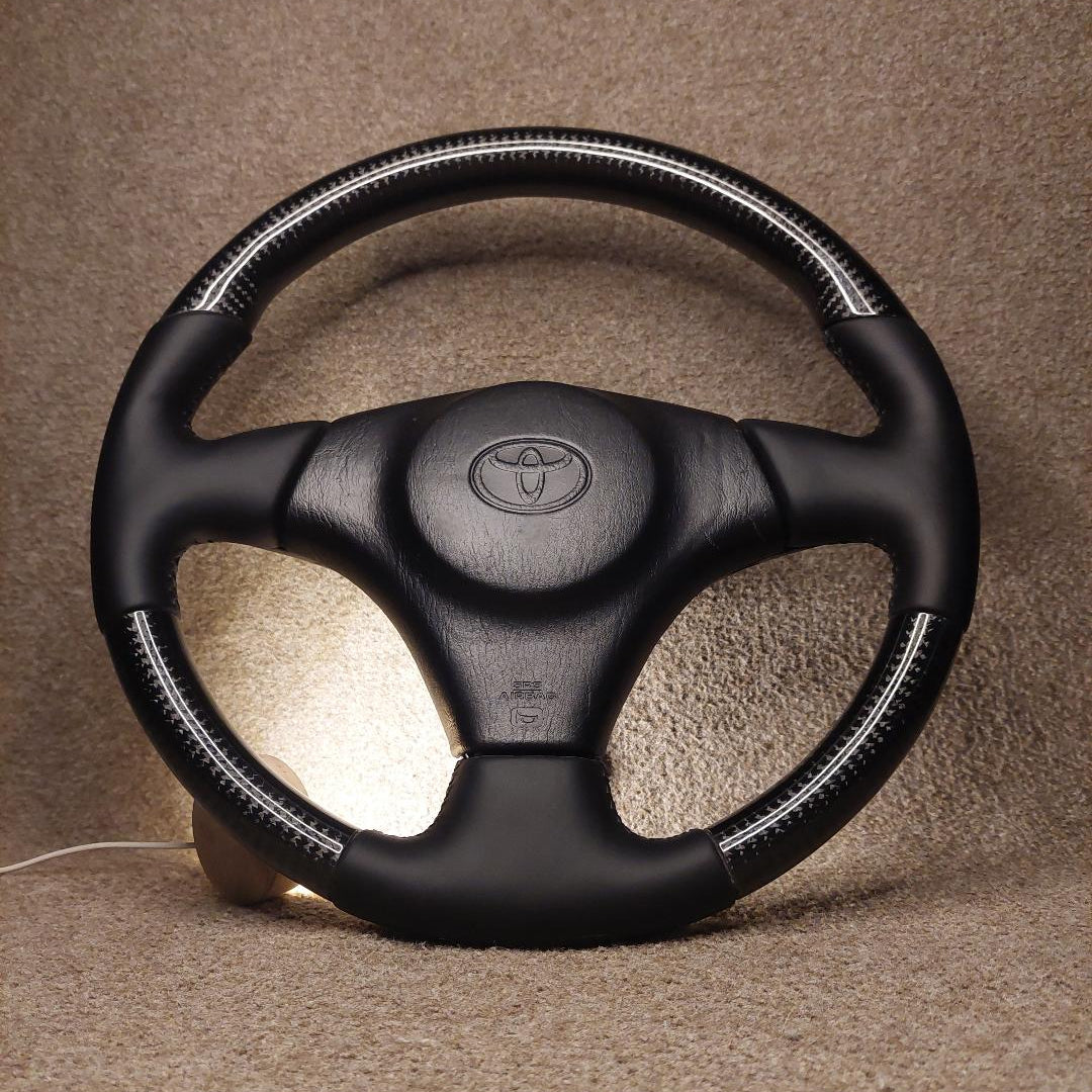 A black leather steering wheel cover with white contrast stitching and a central Toyota emblem, shown on a steering wheel against a textured beige background.