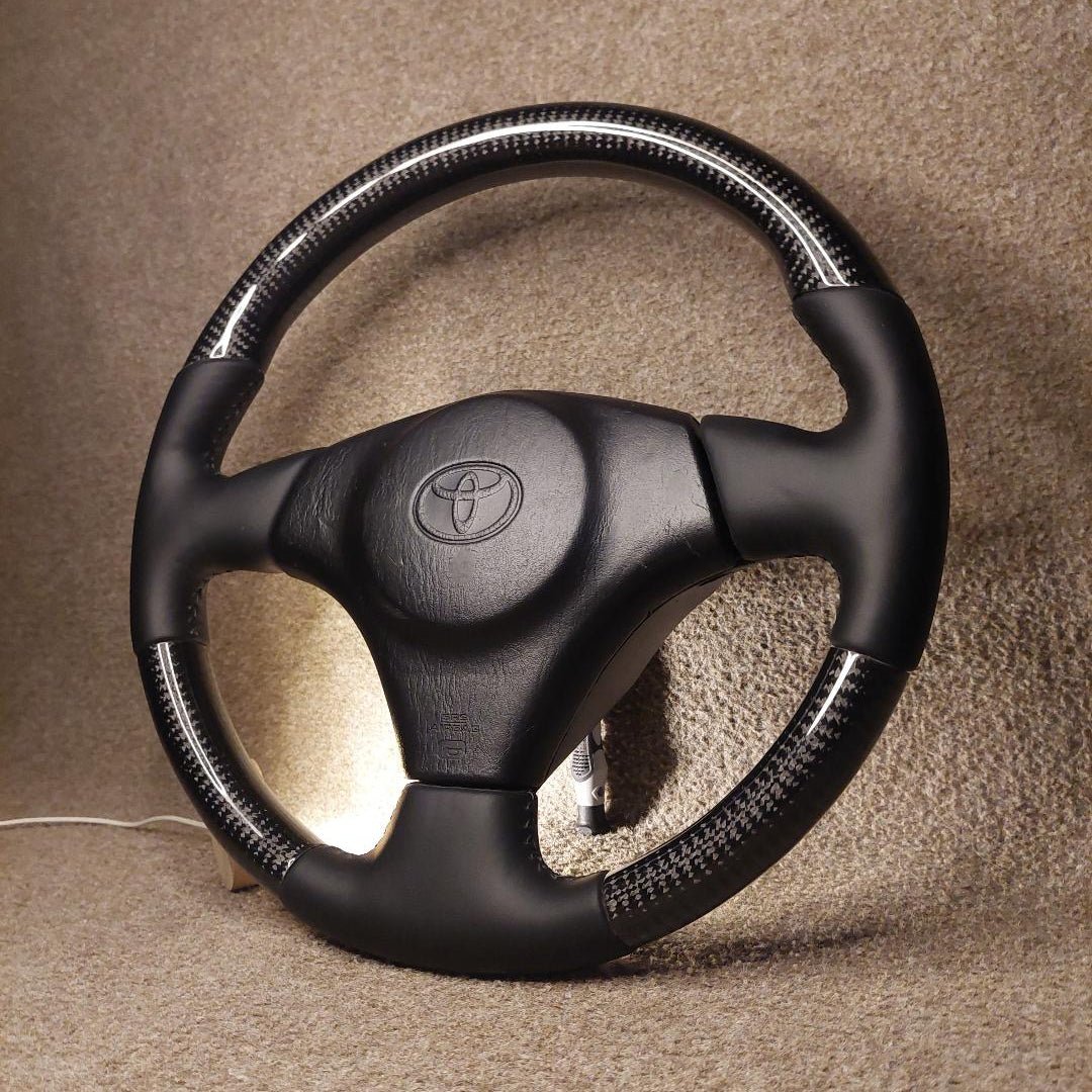 A black steering wheel cover with a carbon fiber pattern wrap around the outer rim, fitted on a Toyota steering wheel. The Toyota emblem is visible in the center.