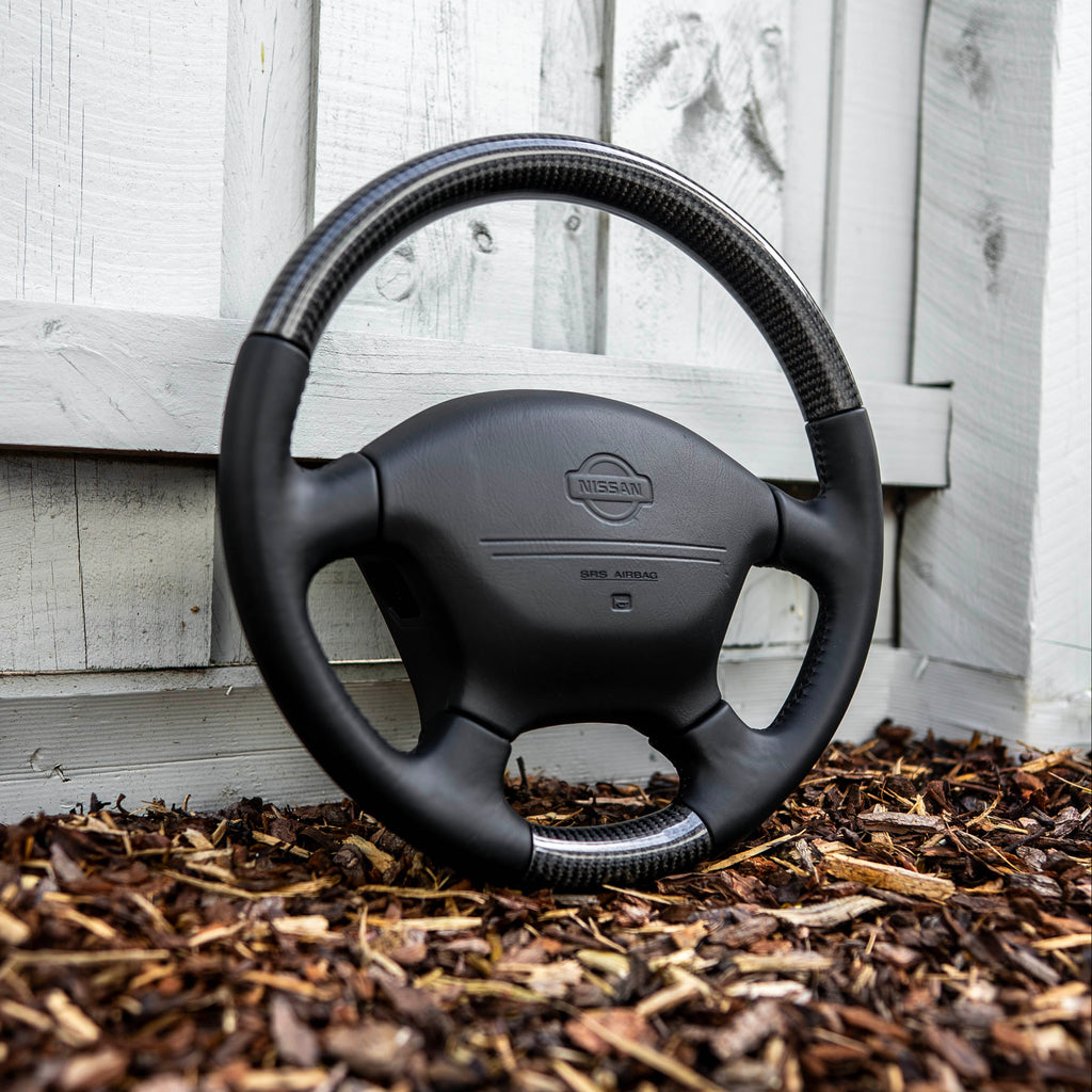 A black steering wheel with a carbon fiber rim wrap and a central Nissan logo, leaning against a white wooden wall on a bed of brown wood mulch.