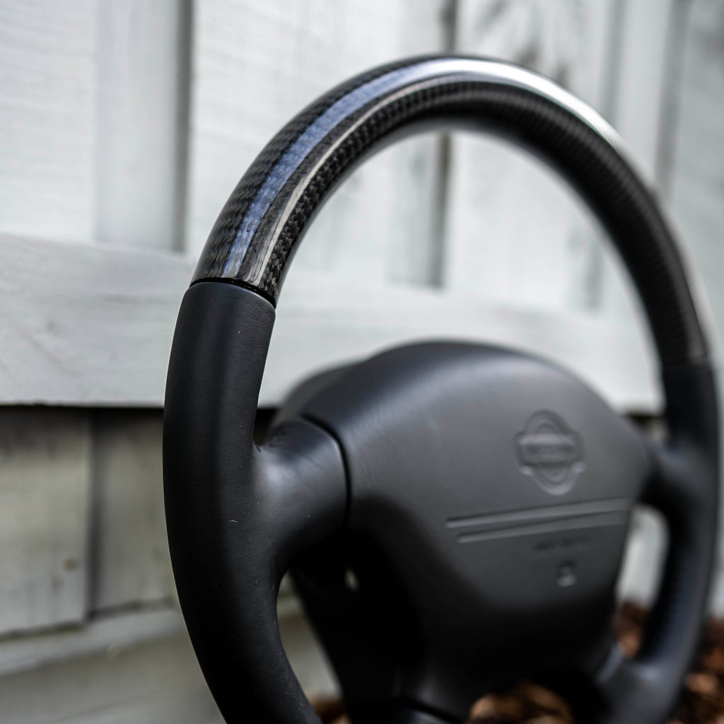 A black carbon fiber steering wheel cover installed on a black steering wheel, leaning against a white wooden fence on a bed of mulch.