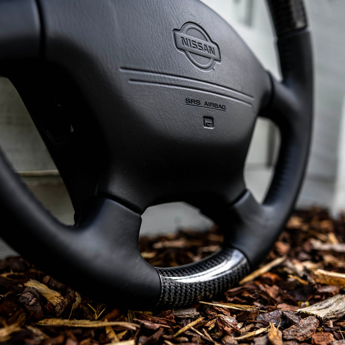 A close-up of a black Nissan steering wheel with a carbon fiber-look lower rim, resting on a bed of brown wood mulch against a light gray wall.